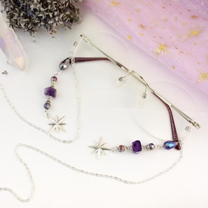 May include: Silver eyeglasses with a chain featuring star charms and purple and iridescent beads. The glasses have a rimless design with a thin silver frame and burgundy temples. The chain is designed to hang around the neck.