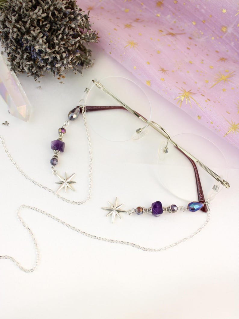 May include: Silver eyeglasses with a chain featuring star charms and purple and iridescent beads. The glasses have a rimless design with a thin silver frame and burgundy temples. The chain is designed to hang around the neck.