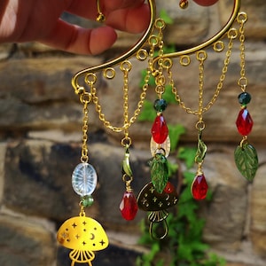 May include: Gold ear cuff with dangling charms, including red and green glass beads, leaves, and a mushroom charm with stars and a crescent moon.