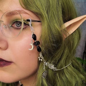 Puede incluir: Una cadena de plata con abalorios, incluyendo una luna creciente, estrellas, un sol y cristales de cuarzo transparentes, cuelga de unas gafas. La cadena la lleva una persona con el pelo verde y orejas de elfo.