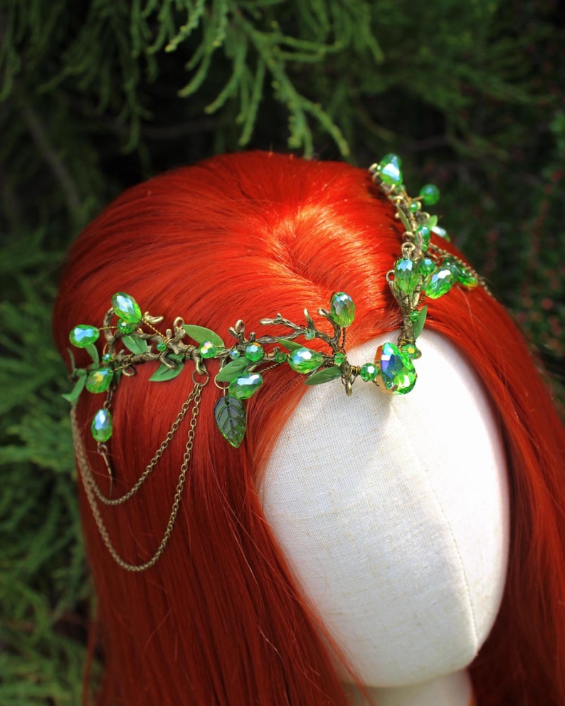 May include: A green and gold-toned headpiece with leaf and crystal accents. The headpiece features a delicate design with green crystals and small leaves. The headpiece is worn on a mannequin head with red hair, set against a green background.