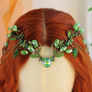 May include: A green and gold headpiece with leaf and crystal accents. The headpiece features a central teardrop-shaped crystal and delicate chain details. The leaves and crystals are in varying shades of green.