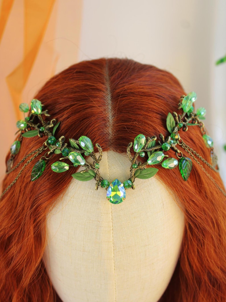May include: A green and gold headpiece with leaf and crystal accents. The headpiece features a central teardrop-shaped crystal and delicate chain details. The leaves and crystals are in varying shades of green.