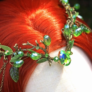 May include: A detailed headpiece with green crystal accents and leaf details. The crown features a bronze-colored metal frame with dangling chains and is set against a backdrop of red hair.