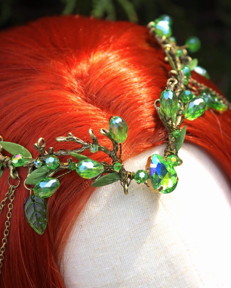 May include: A detailed headpiece with green crystal accents and leaf details. The crown features a bronze-colored metal frame with dangling chains and is set against a backdrop of red hair.
