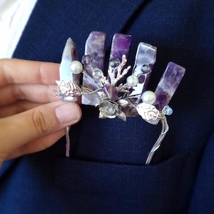 May include: A lapel pin with amethyst crystals and silver wire. The pin features a cluster of purple crystals, pearls, and silver leaves. The pin is set against a navy blue jacket, a stylish accessory.
