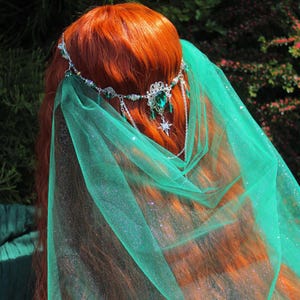May include: A silver-toned headpiece featuring a central emerald green teardrop gemstone, intricate metal filigree, and a dangling star charm. The headpiece is adorned with small green beads and is paired with a sheer, shimmering green tulle veil.