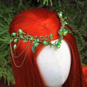 May include: A decorative headpiece with green gemstones and leaves. The headpiece is made of gold-colored metal and features dangling chains. The headpiece is displayed on a mannequin head with red hair.