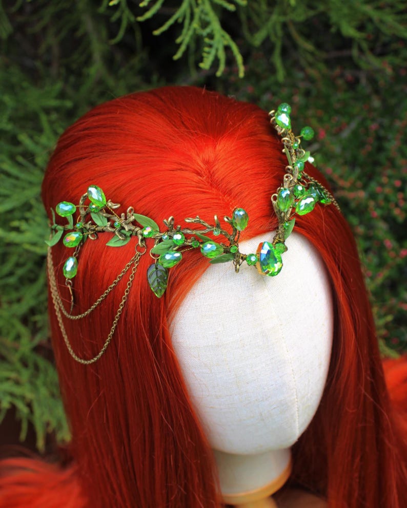 May include: A decorative headpiece with green gemstones and leaves. The headpiece is made of gold-colored metal and features dangling chains. The headpiece is displayed on a mannequin head with red hair.
