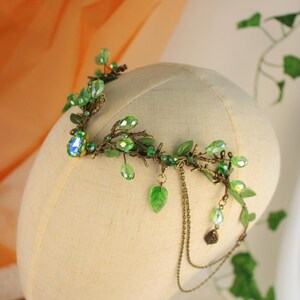 May include: A handcrafted headpiece featuring a botanical design. The crown is adorned with green leaves, sparkling green beads, and a large, iridescent gemstone. Delicate chains hang from the side, adding movement. The headpiece is displayed on a mannequin head.