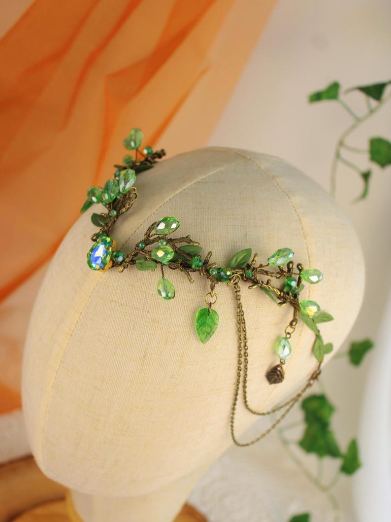 May include: A handcrafted headpiece featuring a botanical design. The crown is adorned with green leaves, sparkling green beads, and a large, iridescent gemstone. Delicate chains hang from the side, adding movement. The headpiece is displayed on a mannequin head.