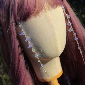 May include: A head mannequin with pink and purple hair, adorned with delicate silver chains. The chains are embellished with small, light purple flower-shaped beads and iridescent crystal beads, creating a whimsical and ethereal aesthetic.