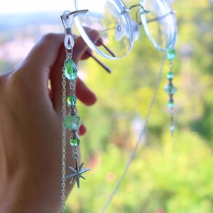 May include: Clear spectacles with a decorative chain. The chain features green beads, a silver star charm, and a delicate silver chain. The background is a blurred view of green foliage.