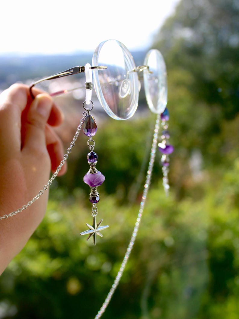 May include: Eyeglass chain with a silver chain and purple beads. The chain is attached to clear eyeglasses. A star charm hangs from the chain. The background is a blurred green.