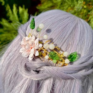 Puede incluir: Un accesorio floral para el cabello con una flor rosa claro, hojas verdes y cuentas blancas. El diseño de alambre dorado está acentuado con cristales transparentes. El adorno para el cabello se coloca sobre un fondo de cabello morado claro y follaje verde.