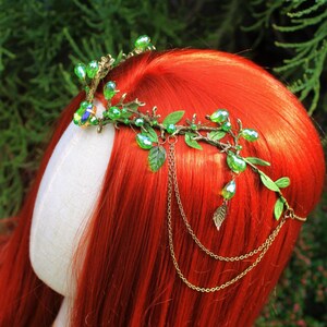 May include: A green and gold headpiece with a botanical design. The headpiece features green crystal beads, small green leaves, and gold-colored chains. The headpiece is displayed on a mannequin head with red hair.