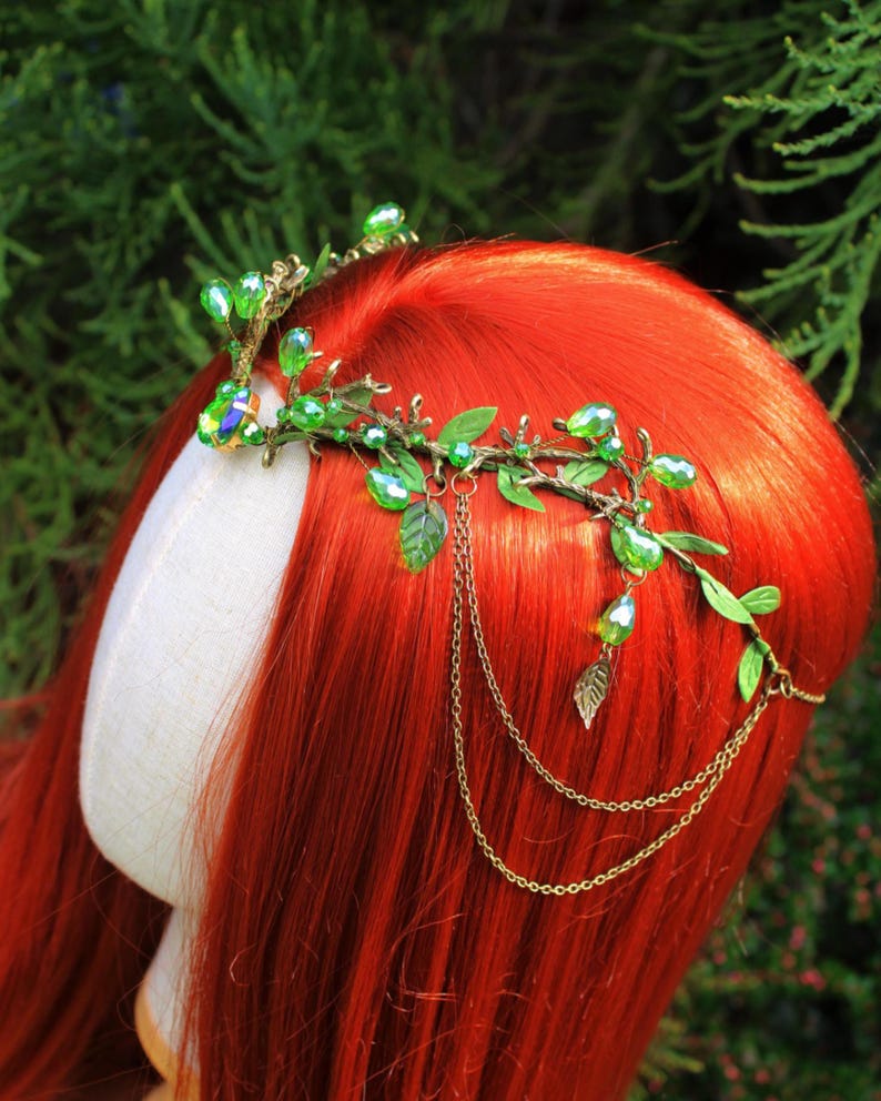 May include: A green and gold headpiece with a botanical design. The headpiece features green crystal beads, small green leaves, and gold-colored chains. The headpiece is displayed on a mannequin head with red hair.