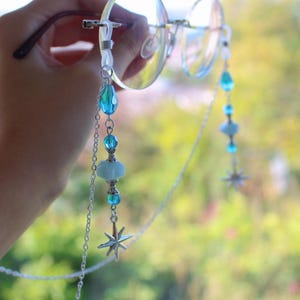 Könnte beinhalten: Brillenkette mit transparenten Gläsern und einer dekorativen Kette. Die Kette hat blaue und türkisfarbene Perlen, einen silbernen Sternanhänger und eine silberne Kette. Die Brille wird von einer Hand gehalten.