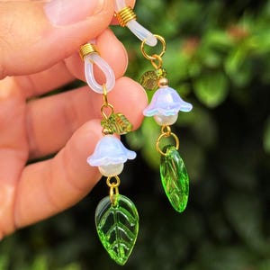 May include: Eyeglass chain with delicate floral and leaf charms. The chain features clear loops, gold-colored metal accents, and dangling charms including light blue flower-shaped beads, green leaf-shaped beads, and small green leaf accents.