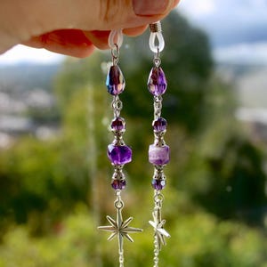 May include: A pair of eyeglass chains with purple and silver beads. The chains feature teardrop-shaped beads, faceted purple beads, and star-shaped charms. The chains are held up by a hand against a blurred green background.