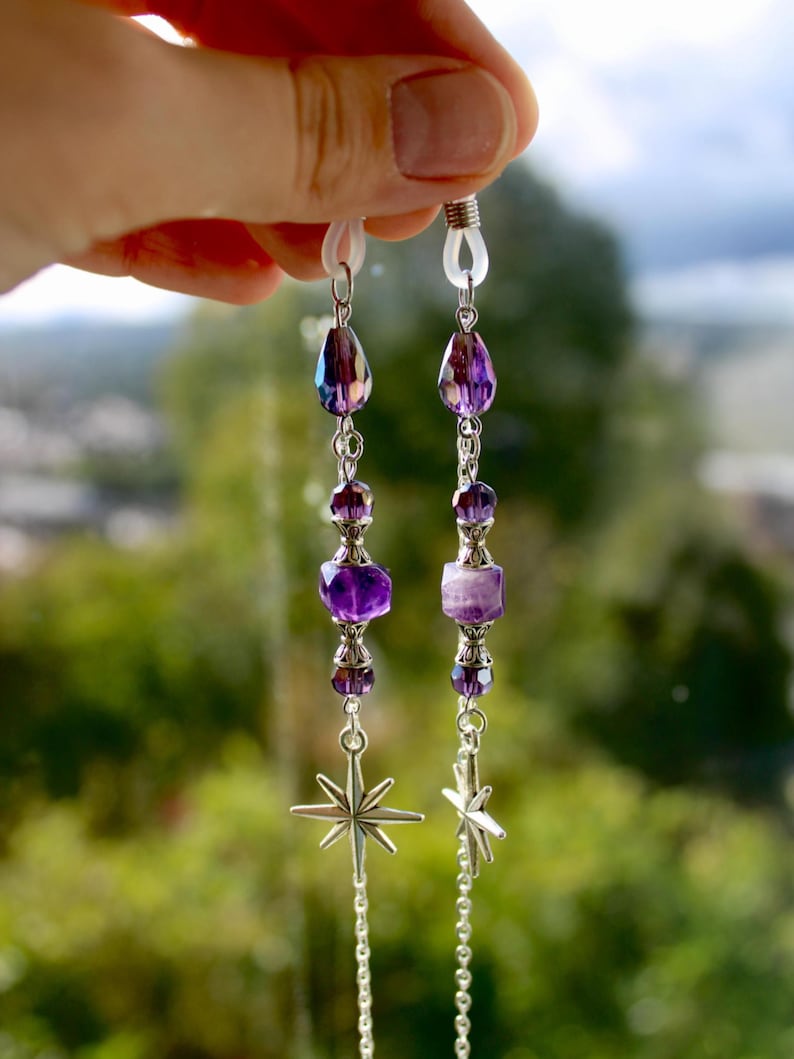 May include: A pair of eyeglass chains with purple and silver beads. The chains feature teardrop-shaped beads, faceted purple beads, and star-shaped charms. The chains are held up by a hand against a blurred green background.