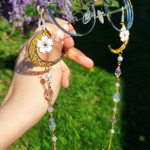 Könnte beinhalten: Eine goldene Kette mit einem Halbmond-Anhänger und einem kleinen weißen Blumen-Anhänger. Die Kette ist mit schillernden Perlen verziert und an einer runden Brille befestigt.