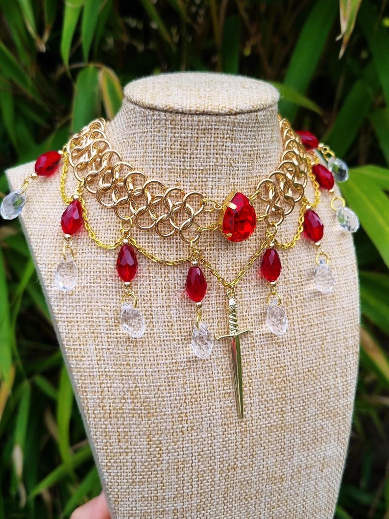 May include: A gold chainmail necklace with red and clear glass beads, a sword pendant, and a large red gemstone in the centre.