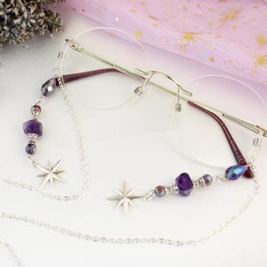 May include: Rimless eyeglasses with a silver chain eyeglass holder. The chain features star charms and purple and silver beads. The glasses have a silver frame and burgundy temples. A sprig of lavender and a crystal are in the background.
