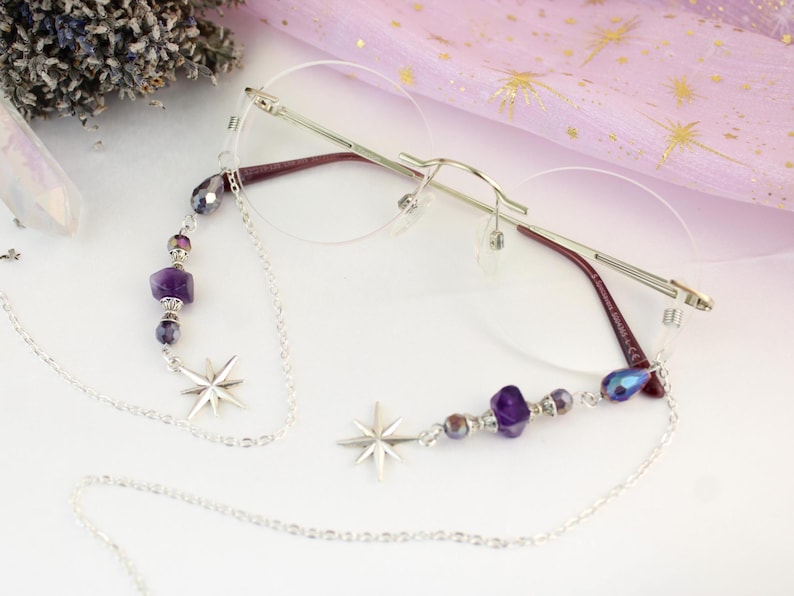 May include: Rimless eyeglasses with a silver chain eyeglass holder. The chain features star charms and purple and silver beads. The glasses have a silver frame and burgundy temples. A sprig of lavender and a crystal are in the background.