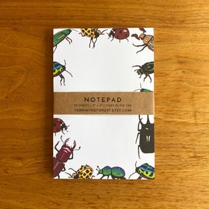 May include: A notepad with a white background and a border of colorful beetles. The notepad has 50 sheets and is 4 x 6 inches. The text "NOTEPAD" is printed on the front of the notepad, along with the text "50 SHEETS | 4" x 6" | MADE IN THE USA" and "FERNINTHEFOREST.ETSY.COM".