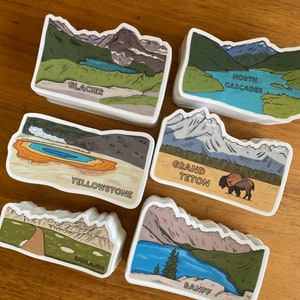 May include: A set of ten illustrated stickers featuring national parks in the United States. The stickers are white with colorful illustrations of mountains, lakes, and other natural features. The names of the parks are printed in black text below each illustration. The parks featured are Glacier, Yellowstone, Grand Teton, Badlands, and Banff.