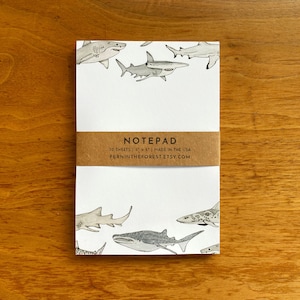 May include: A white notepad with a shark illustration. The notepad is wrapped with a brown paper band that reads "NOTEPAD" and "FERNINTHEFOREST.ETSY.COM". The notepad measures 4 x 6 inches and contains 50 sheets.