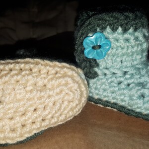 May include: Handmade crochet baby booties in light blue and cream colors. One bootie is upright, showing a turquoise flower button. The other bootie is lying down. The booties have a dark green trim.