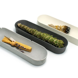May include: Three oval-shaped ceramic dishes in grey, black and white, each containing a different type of smudge stick. The grey dish holds a palo santo stick, the black dish holds a bundle of sage, and the white dish holds a white sage smudge stick.