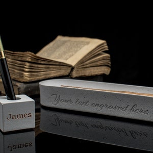 Puede incluir: Un portaplumas de hormig&oacute;n gris con el nombre "James" grabado en la parte delantera. Un bol&iacute;grafo dorado y negro est&aacute; en el portaplumas. Una bandeja de hormig&oacute;n gris con el texto "Your text engraved here" est&aacute; en el fondo.