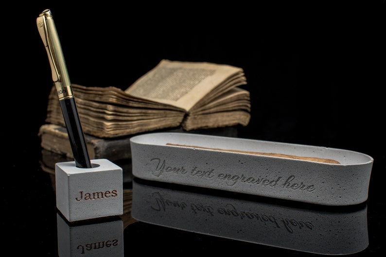 Puede incluir: Un portaplumas de hormig&oacute;n gris con el nombre "James" grabado en la parte delantera. Un bol&iacute;grafo dorado y negro est&aacute; en el portaplumas. Una bandeja de hormig&oacute;n gris con el texto "Your text engraved here" est&aacute; en el fondo.