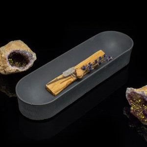 Puede incluir: Un plato ovalado de cer&aacute;mica gris que contiene un palo santo y flores de lavanda secas. Dos geodas de amatista con cristales dorados est&aacute;n a cada lado del plato.