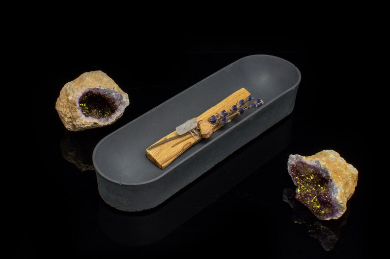 Puede incluir: Un plato ovalado de cer&aacute;mica gris que contiene un palo santo y flores de lavanda secas. Dos geodas de amatista con cristales dorados est&aacute;n a cada lado del plato.