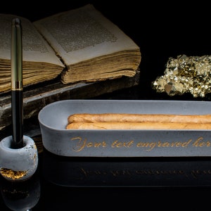 Puede incluir: Un cenicero de cigarros de hormig&oacute;n gris con dos cigarros dentro. El cenicero tiene las palabras "Your text engraved here" escritas en letras doradas. Un portaplumas con una pluma dorada est&aacute; al lado del cenicero. Un mont&oacute;n de libros antiguos y un grupo de cristales de pirita dorada est&aacute;n en el fondo.