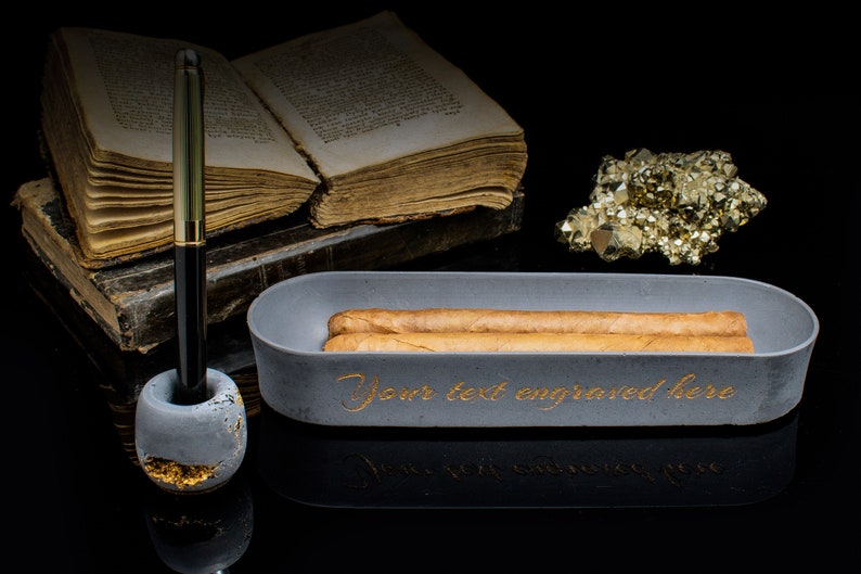 Puede incluir: Un cenicero de cigarros de hormig&oacute;n gris con dos cigarros dentro. El cenicero tiene las palabras "Your text engraved here" escritas en letras doradas. Un portaplumas con una pluma dorada est&aacute; al lado del cenicero. Un mont&oacute;n de libros antiguos y un grupo de cristales de pirita dorada est&aacute;n en el fondo.