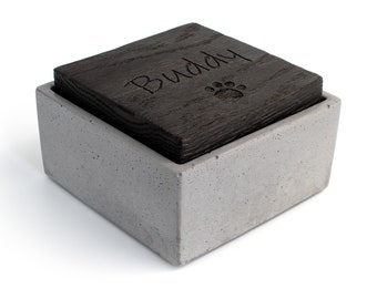 Op de afbeelding: Een grijze betonnen doos met een donkerhouten deksel. Het deksel heeft de naam "Buddy" gegraveerd met een pootprint.