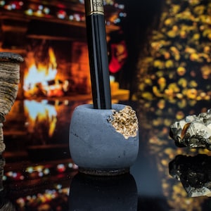 Puede incluir: Un portaplumas de hormig&oacute;n gris con un acento dorado. El portaplumas est&aacute; sobre una superficie negra delante de una chimenea y un &aacute;rbol de Navidad.
