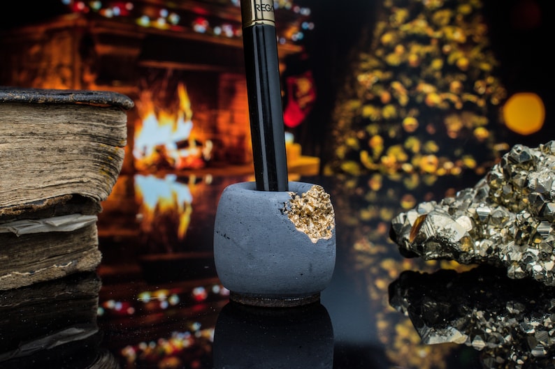 Puede incluir: Un portaplumas de hormig&oacute;n gris con un acento dorado. El portaplumas est&aacute; sobre una superficie negra delante de una chimenea y un &aacute;rbol de Navidad.