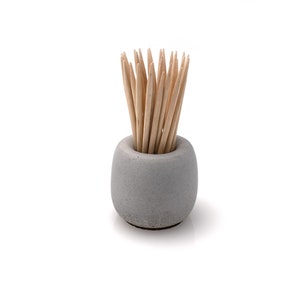 May include: A grey concrete toothpick holder with a round base and wooden toothpicks sticking out of the top.
