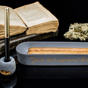 Puede incluir: Un cenicero de cigarros de hormig&oacute;n gris con dos cigarros dentro. El cenicero tiene las palabras "Your text engraved here" escritas en letras doradas. El cenicero est&aacute; sobre una superficie negra junto a un libro, un portaplumas y un mont&oacute;n de pirita dorada.
