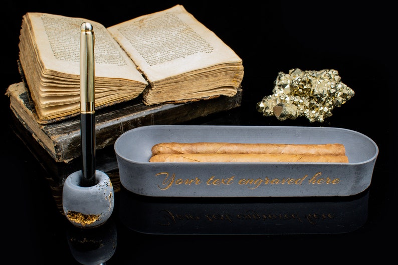 Puede incluir: Un cenicero de cigarros de hormig&oacute;n gris con dos cigarros dentro. El cenicero tiene las palabras "Your text engraved here" escritas en letras doradas. El cenicero est&aacute; sobre una superficie negra junto a un libro, un portaplumas y un mont&oacute;n de pirita dorada.