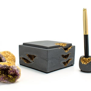 Puede incluir: Una caja de hormig&oacute;n gris con detalles dorados y un portaplumas a juego. La caja tiene tapa y est&aacute; decorada con un patr&oacute;n de escamas doradas. El portaplumas es redondo y tambi&eacute;n est&aacute; decorado con escamas doradas. La caja y el portaplumas est&aacute;n sobre una superficie blanca.
