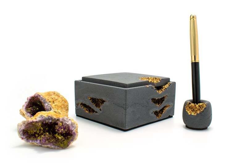 Puede incluir: Una caja de hormig&oacute;n gris con detalles dorados y un portaplumas a juego. La caja tiene tapa y est&aacute; decorada con un patr&oacute;n de escamas doradas. El portaplumas es redondo y tambi&eacute;n est&aacute; decorado con escamas doradas. La caja y el portaplumas est&aacute;n sobre una superficie blanca.
