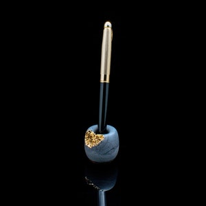 Puede incluir: Un bol&iacute;grafo negro y dorado en un porta bol&iacute;grafos de cer&aacute;mica gris con detalles dorados.