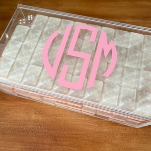 Op de afbeelding: Een doorzichtige acrylbox met een roze monogram op het deksel. De doos is gevuld met kleine, vierkante, parelmoerachtige objecten. De doos staat op een houten oppervlak.